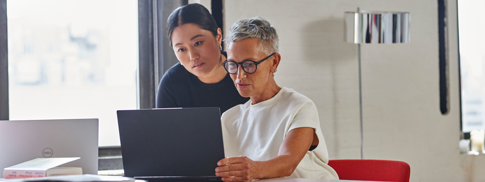 A standing woman leans over the shoulder of a seated women, as both stare at the screen of an open laptop.