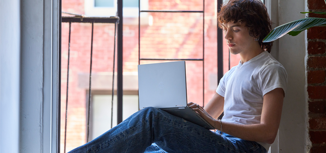 A young person sitting in the windowsill typing on a laptop.