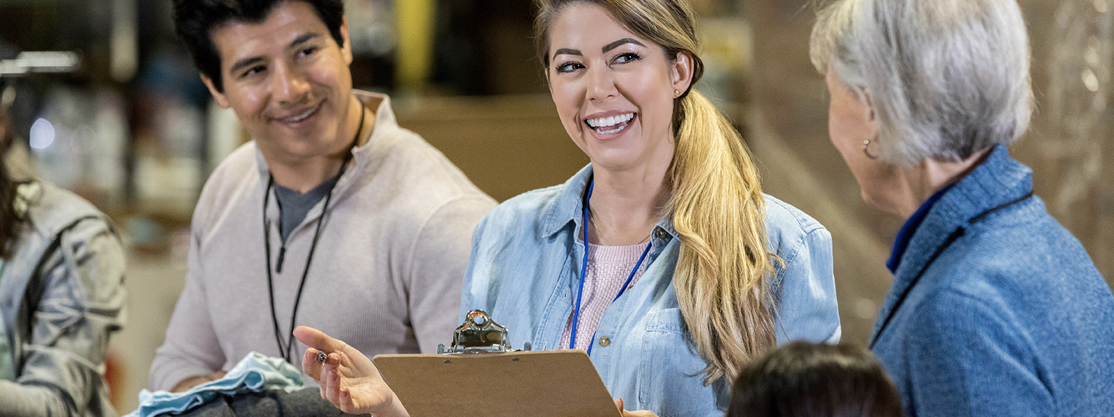 A smiling woman stands in a food bank warehouse and holds a clipboard. She is discussing procedure with volunteers.