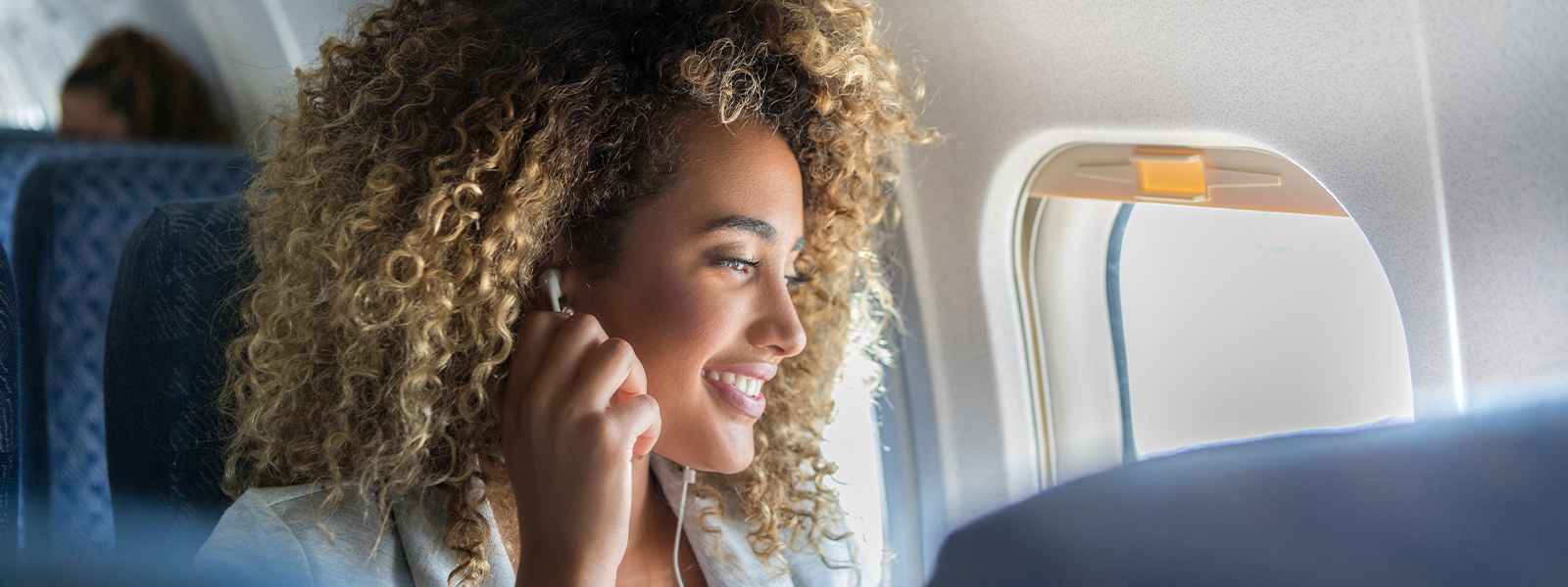 A young woman with curly hair sits in a window seat on a plane. She has her earbuds in as she looks out of the window and smiles.