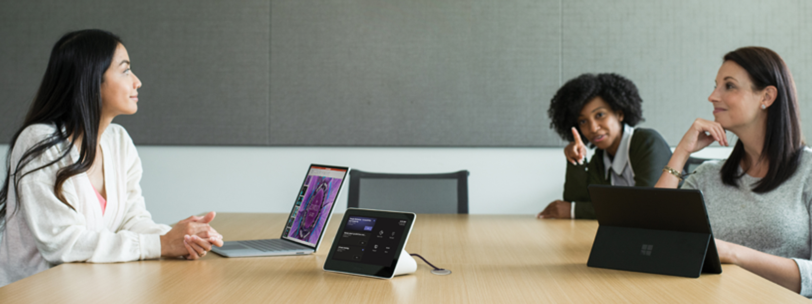 Three females in a medium conference room featuring an Poly Teams Meeting Rooms touch display with the Teams Meeting pre-join screen in view. 
