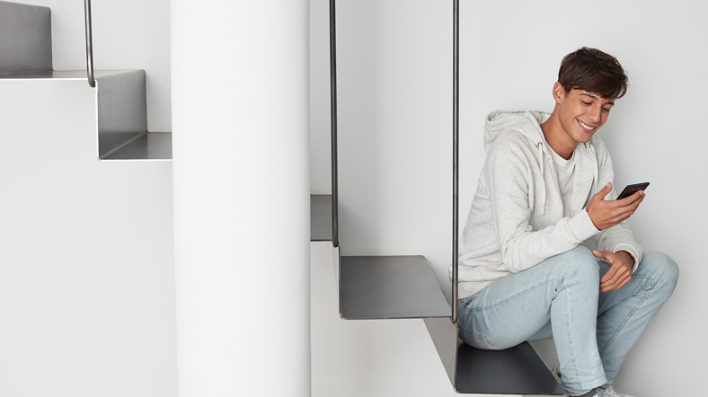 a male student inside home, smiling, and looking at his phone.